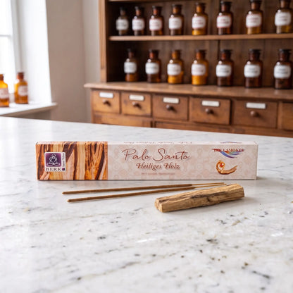 Räucherstäbchen Palo Santo von Berk: Packung auf einer Marmortheke vor einem Apothekerschrank, davor zwei lose Räucherstäbchen und ein Stück Palo-Santo-Holz – Spirit of Gaia
