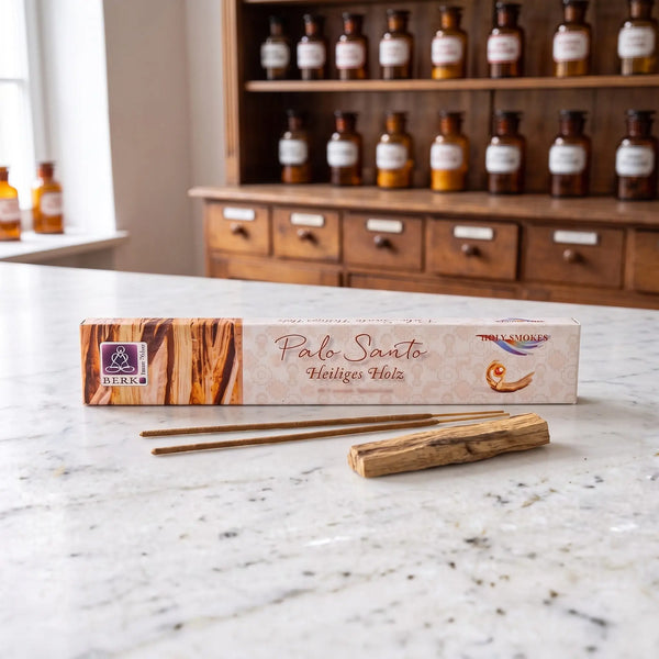 Räucherstäbchen Palo Santo von Berk: Packung auf einer Marmortheke vor einem Apothekerschrank, davor zwei lose Räucherstäbchen und ein Stück Palo-Santo-Holz – Spirit of Gaia