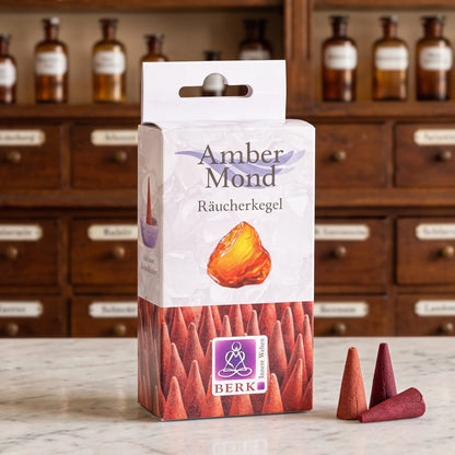 Amber Mond Räucherkegel von Berk: Packung auf einer Marmortheke vor einem Apothekerschrank, daneben drei lose ziegelrote Räucherkegel – Spirit of Gaia