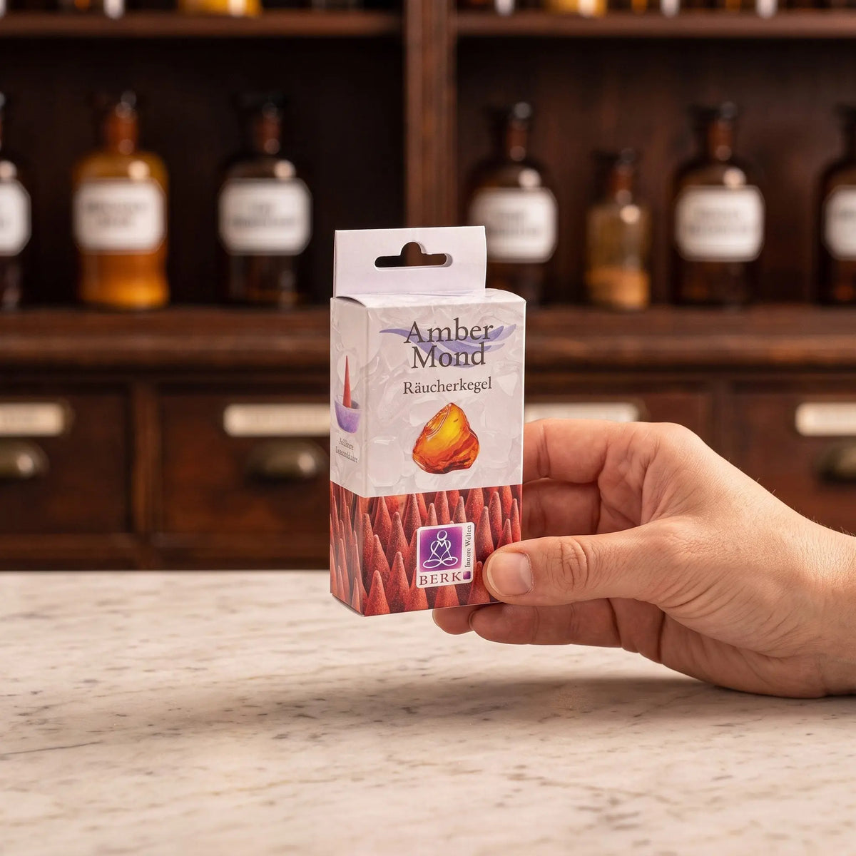Größenvergleich: eine Hand hält die schmale Packung der Amber Mond Räucherkegel von Berk vor dem Apothekerschrank