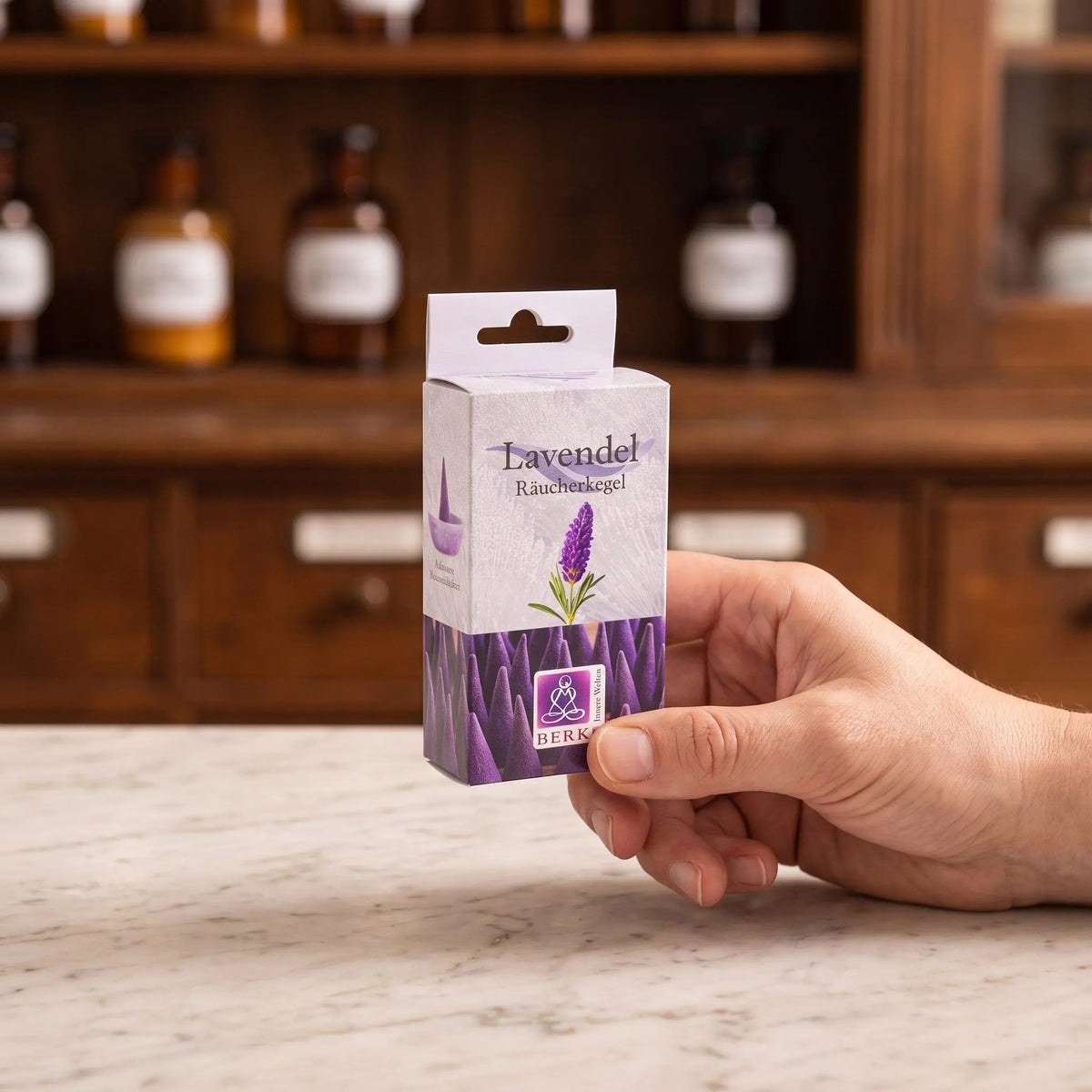 Größenvergleich: eine Hand hält die schmale Packung der Lavendelblüten Räucherkegel von Berk vor dem Apothekerschrank