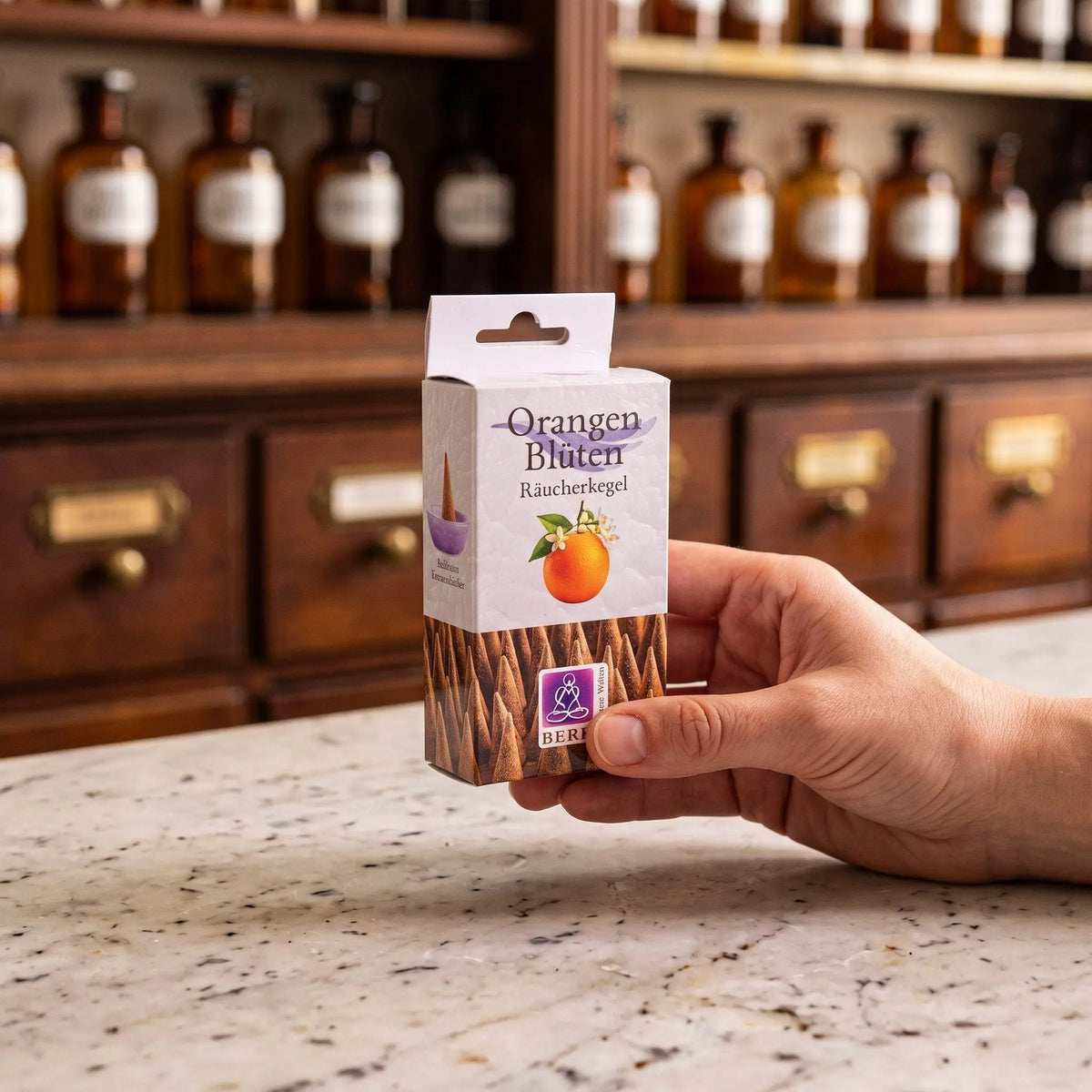 Größenvergleich: eine Hand hält die schmale Packung der Orangenblüten Räucherkegel von Berk vor dem Apothekerschrank