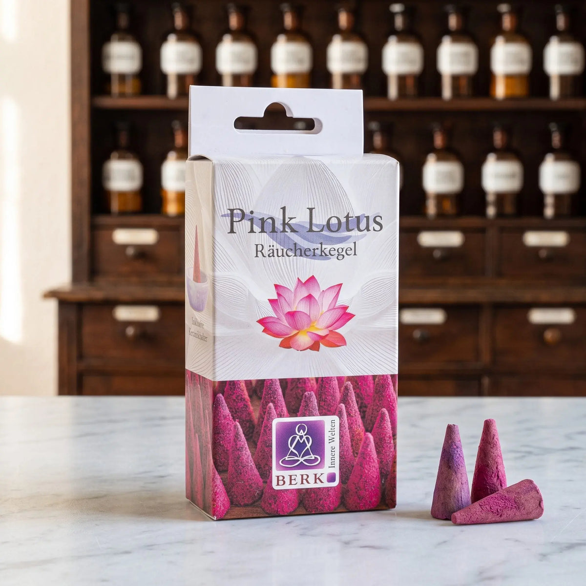 Pink Lotus Räucherkegel von Berk: Packung auf einer Marmortheke vor einem Apothekerschrank, daneben drei lose beerenrosa Räucherkegel – Spirit of Gaia