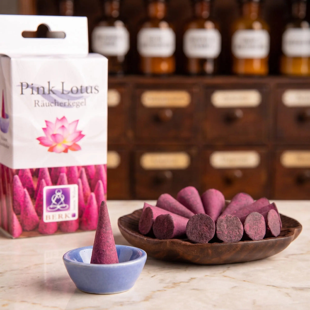 Lieferumfang der Pink Lotus Räucherkegel von Berk: beerenrosa Kegel in einer Holzschale, ein Kegel im beiliegenden blauen Keramikschälchen, dahinter die Packung
