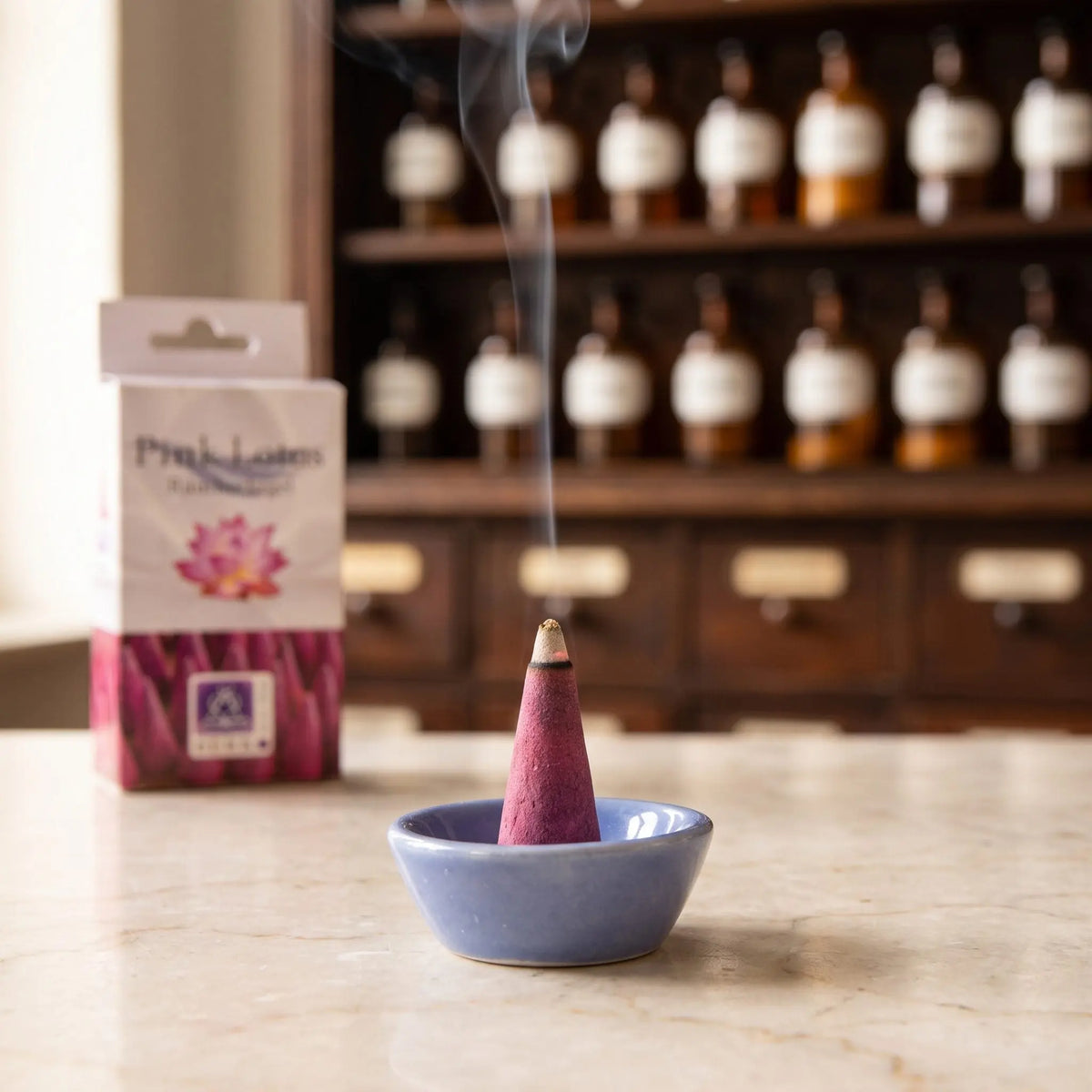 Ein Pink Lotus Räucherkegel glimmt im beiliegenden Keramikschälchen, ein feiner Rauchfaden steigt senkrecht auf