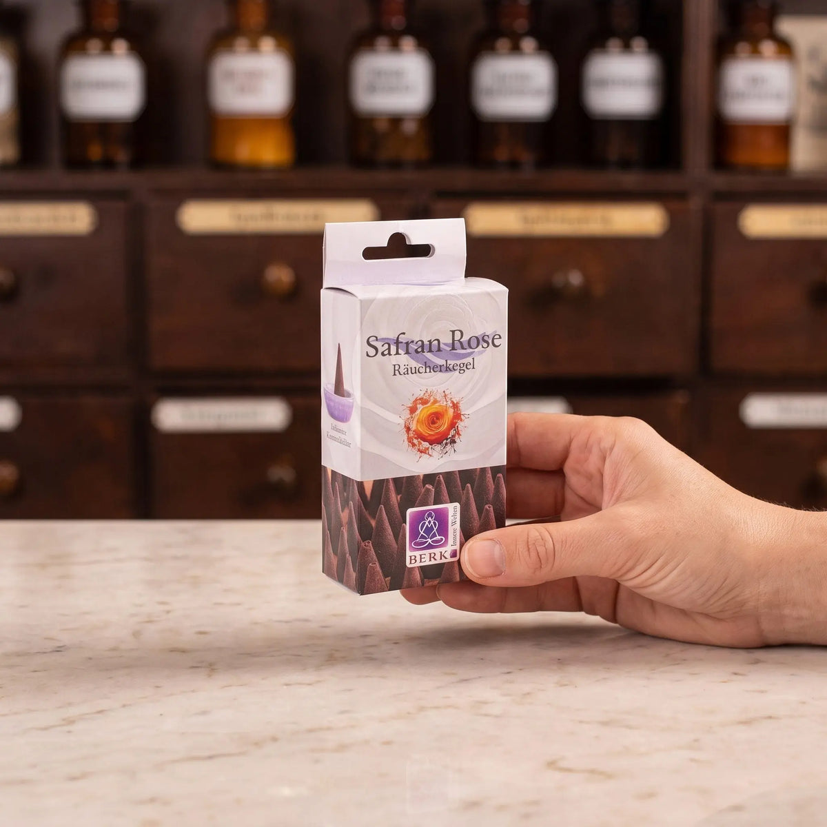 Größenvergleich: eine Hand hält die schmale Packung der Safran Rose Räucherkegel von Berk vor dem Apothekerschrank