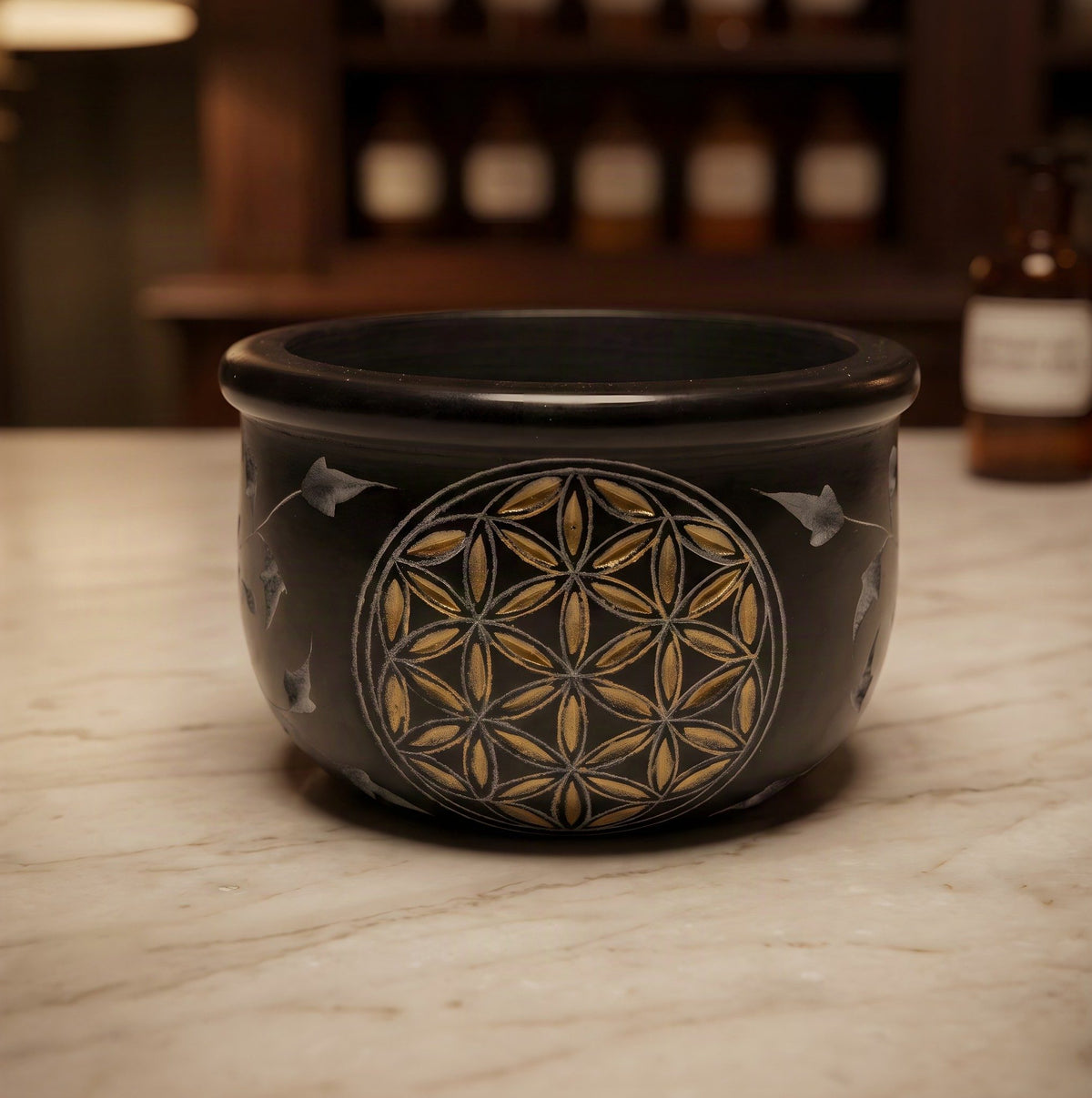 Räucherschale Blume des Lebens von Berk: kleine schwarze Specksteinschale mit goldener Gravur und Blattranken auf der Marmortheke einer Apotheke am Abend