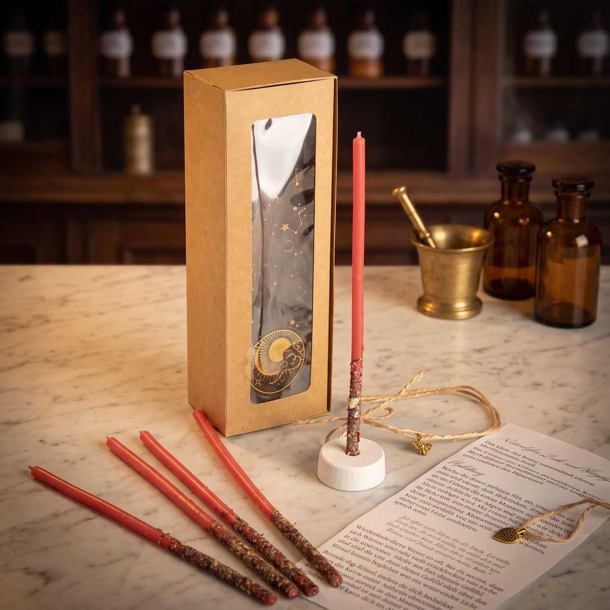 Ritualkerzenset Herzflamme: Kraftpapierschachtel mit Sichtfenster, fünf rote Bienenwachskerzen, Kerzenständer, Herz-Amulett und gerollte Anleitung auf der Marmortheke einer Apotheke am Abend