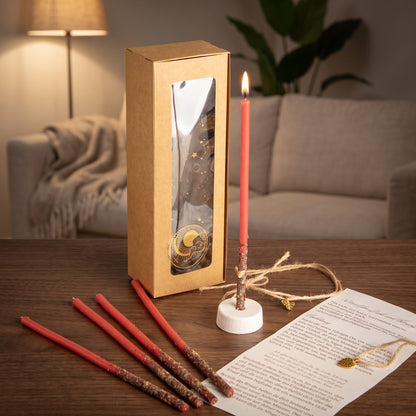 Das Set Herzflamme auf einem dunklen Holztisch im Wohnzimmer am Abend, eine Kerze brennt im Ständer, vier weitere liegen davor, dazu die Anleitung und das Amulett