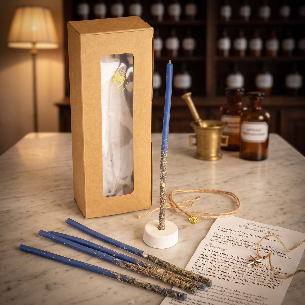 Ritualkerzenset Licht der Wahrheit: Kraftpapierschachtel mit Sichtfenster, fünf blaue Bienenwachskerzen, Kerzenständer, Leitstern-Amulett und gerollte Anleitung auf der Marmortheke einer Apotheke am Abend
