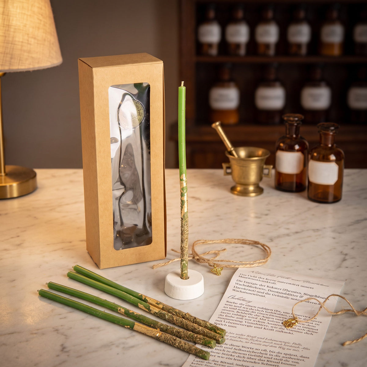 Ritualkerzenset Wachstumslicht: Kraftpapierschachtel mit Sichtfenster, fünf grüne Bienenwachskerzen, Kerzenständer, Kleeblatt-Amulett und gerollte Anleitung auf der Marmortheke einer Apotheke am Abend