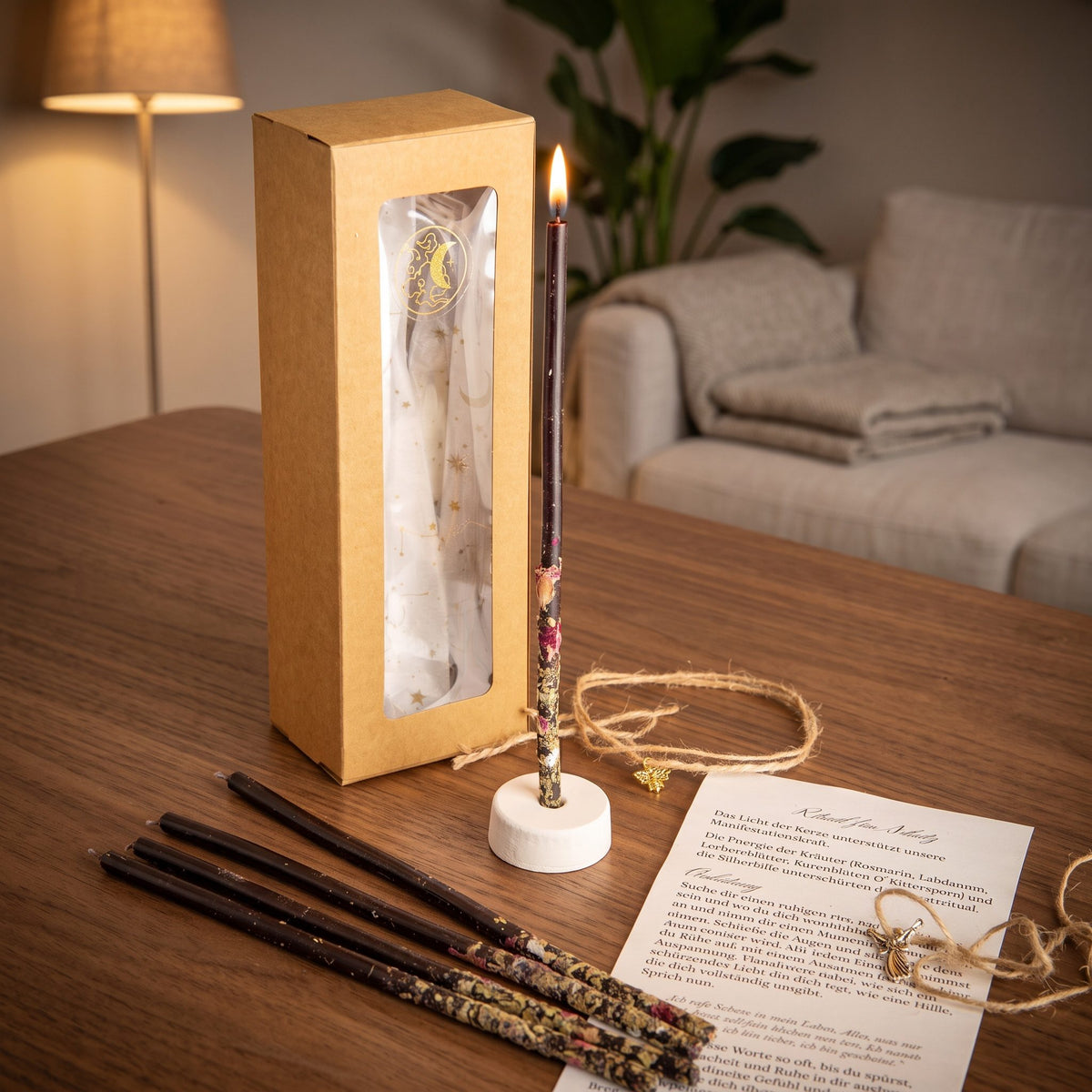 Das Set Wächterlicht auf einem dunklen Holztisch im Wohnzimmer am Abend, eine Kerze brennt im Ständer, vier weitere liegen davor, dazu die Anleitung und das Amulett