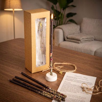 Das Set Wächterlicht auf einem dunklen Holztisch im Wohnzimmer am Abend, eine Kerze brennt im Ständer, vier weitere liegen davor, dazu die Anleitung und das Amulett