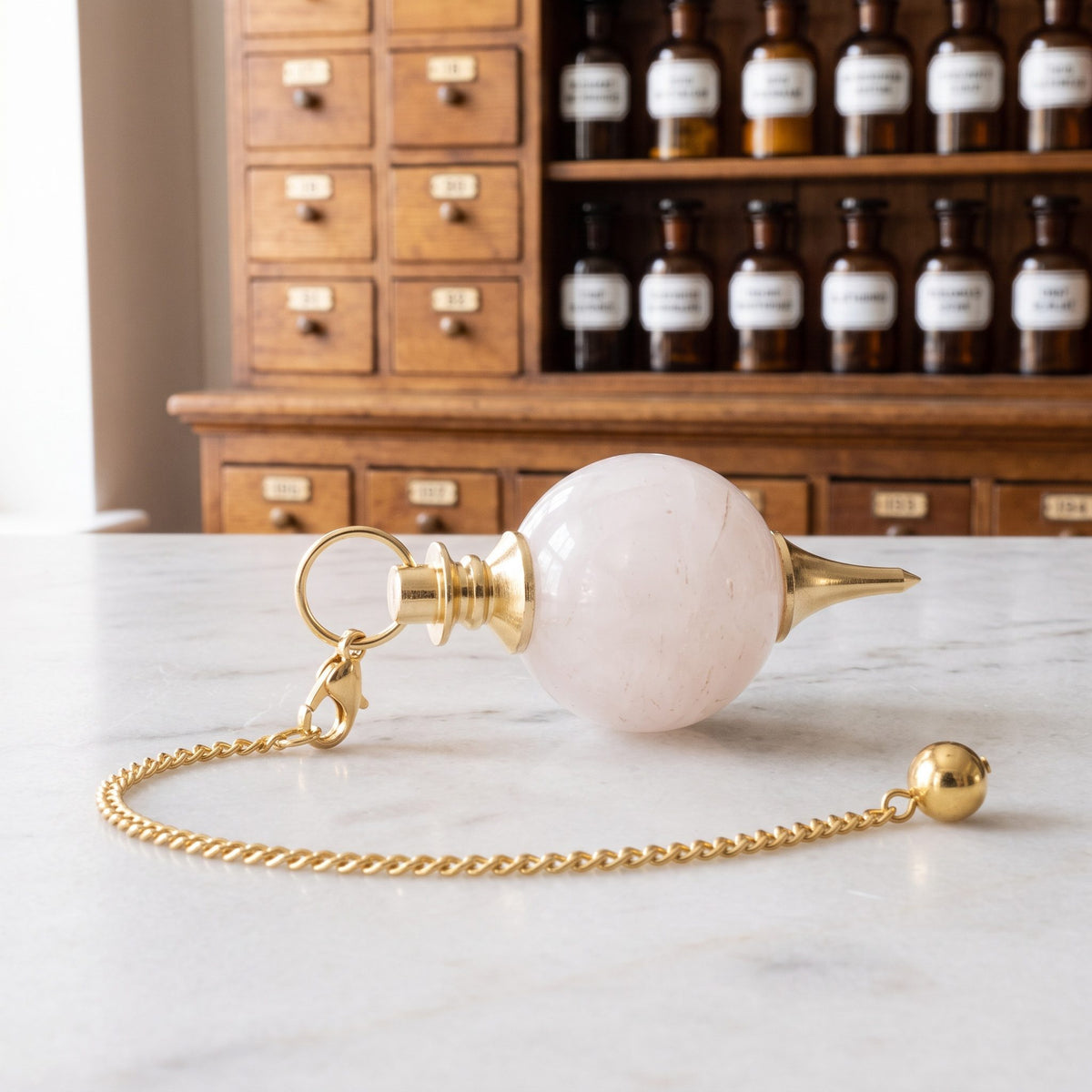 Rosenquarz-Kugelpendel von Berk liegt auf der Marmortheke einer hellen Apotheke, die goldfarbene Kette mit Karabiner und kleiner Endkugel locker daneben