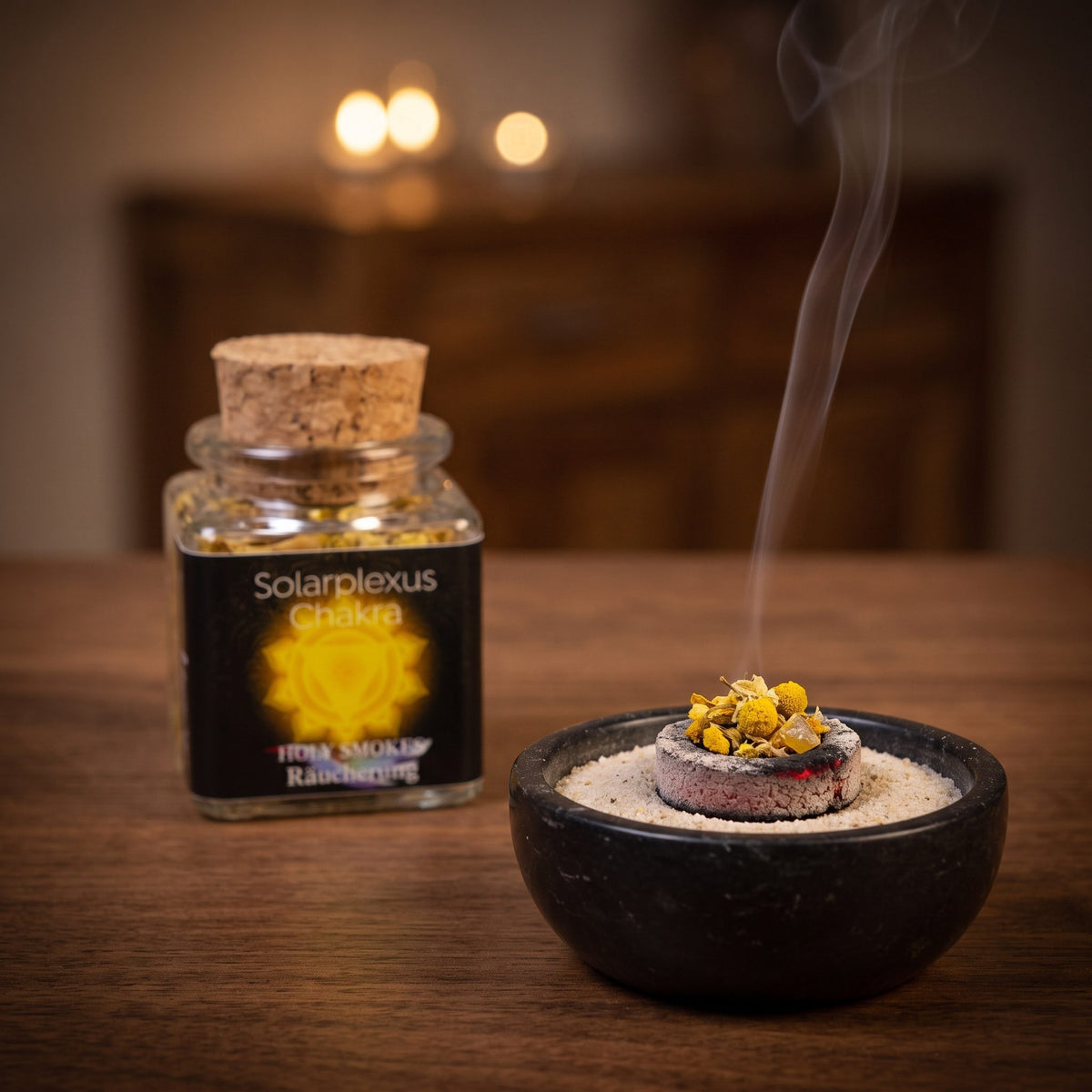 Solarplexus-Chakra, Chakra-Räuchermischung von Berk, 50 ml im Korkglas: glimmende Räucherkohle mit einer Prise der gelben Blütenmischung in einer Specksteinschale, dahinter das Korkglas, feiner Rauch steigt auf einer dunklen Holzplatte am Abend auf – Spirit of Gaia