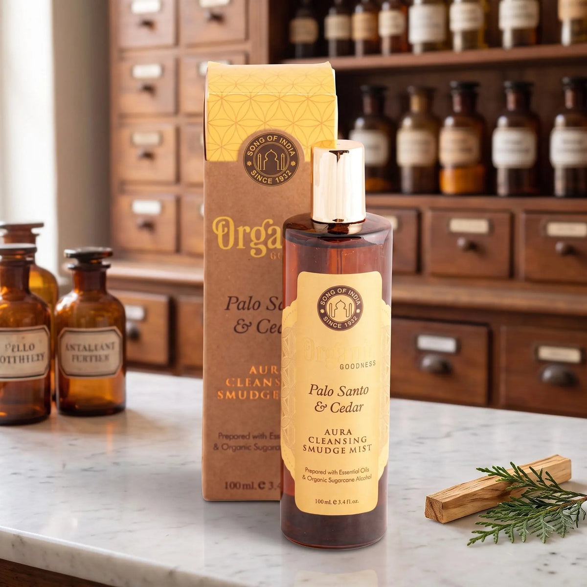 Aura Reinigungsspray Palo Santo und Zeder von Organic Goodness: braune Sprühflasche mit goldener Kappe neben der Kraftpapier-Schachtel auf einer Marmortheke vor einem Apothekerschrank