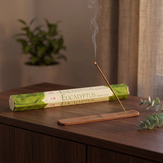 GR Eucalyptus Räucherstäbchen: glimmendes Räucherstäbchen mit feinem Rauch im Holzhalter, dahinter die Sechseck-Packung, auf einem Sideboard am Abend