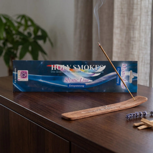 Entspannung Räucherstäbchen Holy Smokes Mystic Line: glimmendes Räucherstäbchen mit feinem Rauch im Holzhalter, dahinter die Packung, auf einem Sideboard am Abend