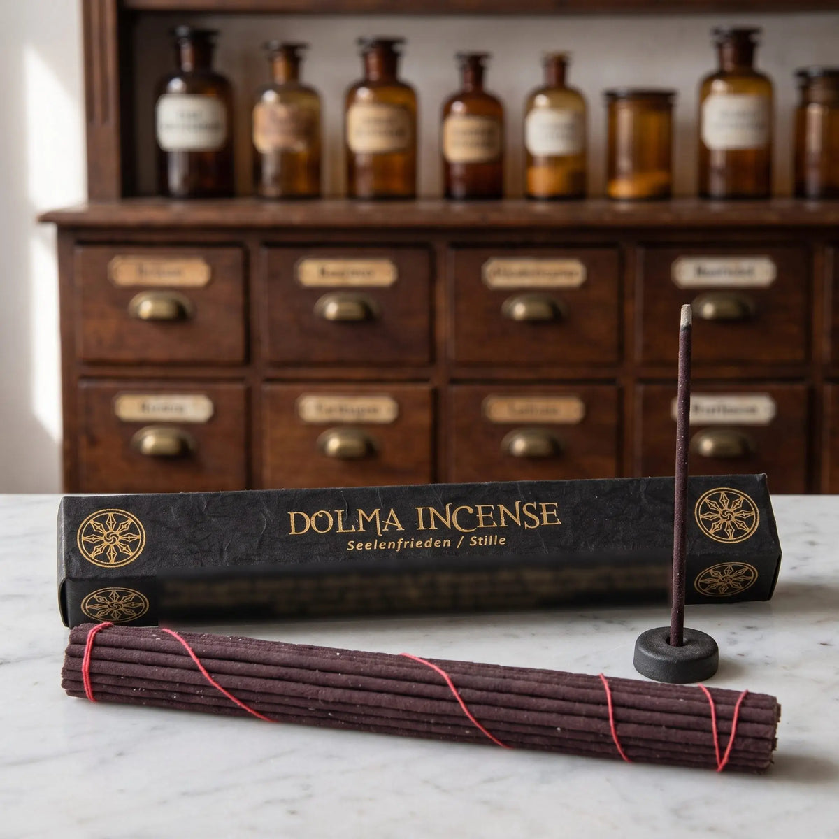 Tibetische Räucherstäbchen Dolma Incense von Berk: schwarze Schachtel mit Goldschrift auf einer Marmortheke vor einem Apothekerschrank, davor das Stäbchenbündel und der Tonhalter