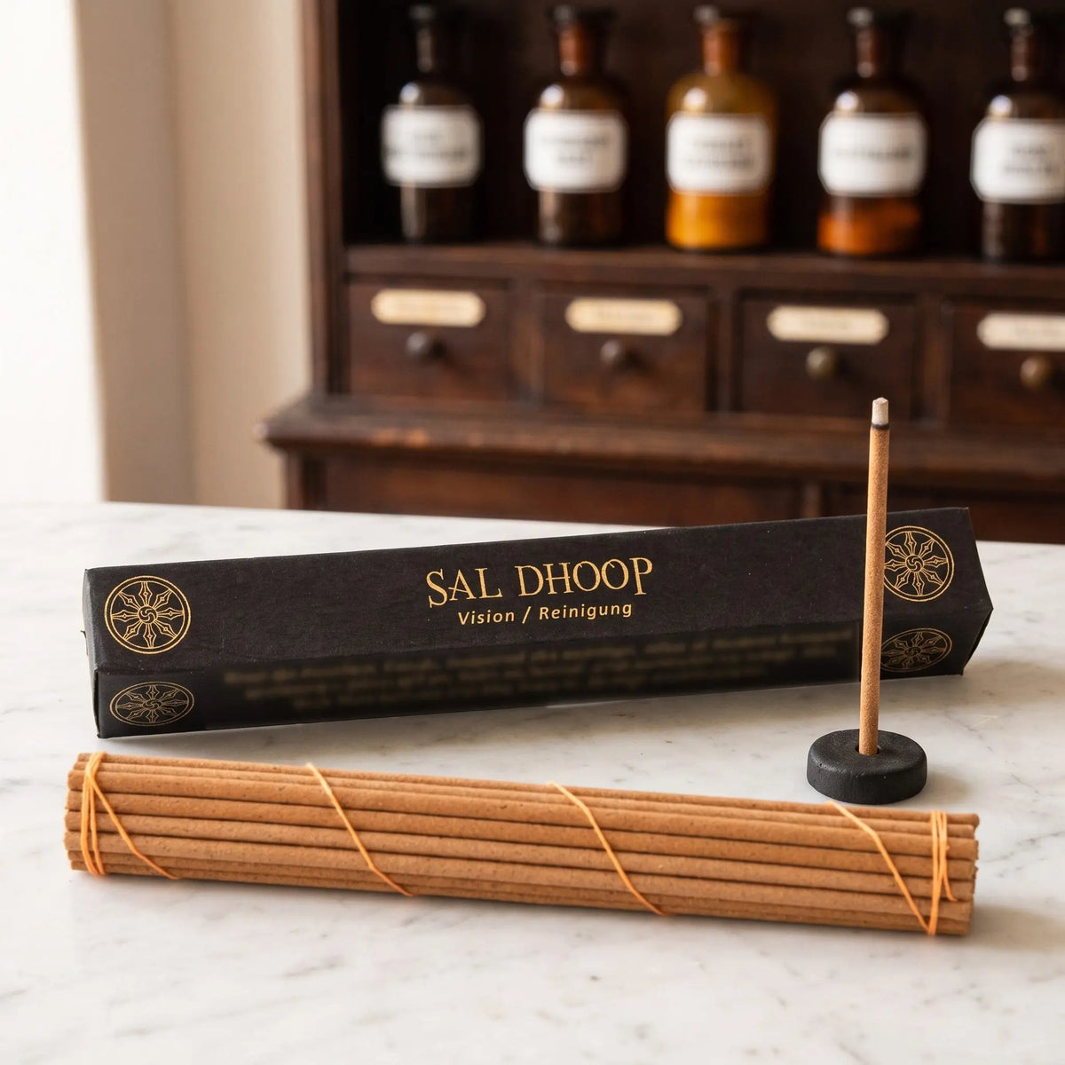 Tibetische Räucherstäbchen Sal Dhoop von Berk: schwarze Schachtel mit Goldschrift auf einer Marmortheke vor einem Apothekerschrank, davor das Stäbchenbündel und der Tonhalter