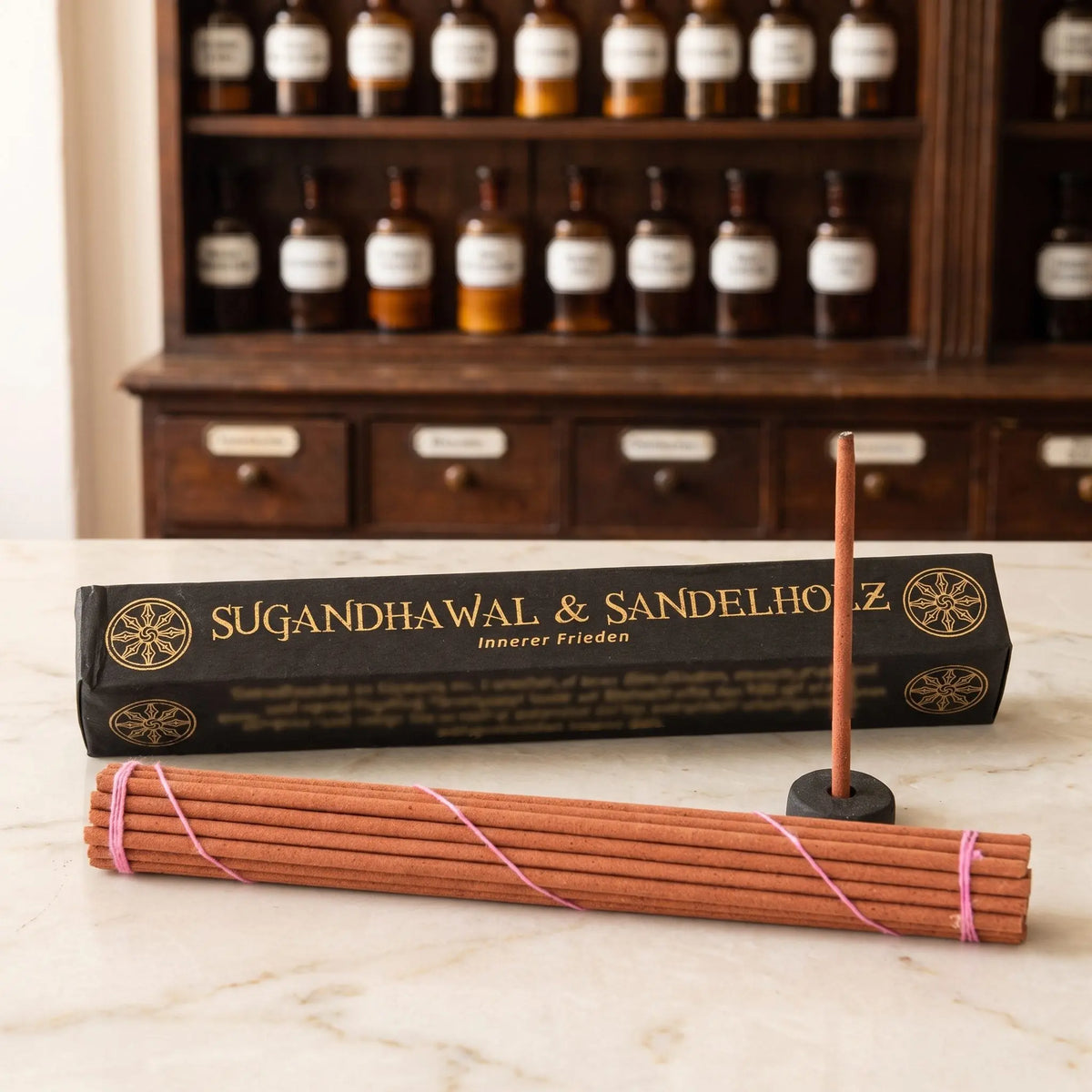 Tibetische Räucherstäbchen Sugandhawal und Sandelholz von Berk: schwarze Schachtel mit Goldschrift auf einer Marmortheke vor einem Apothekerschrank, davor das Stäbchenbündel und der Tonhalter