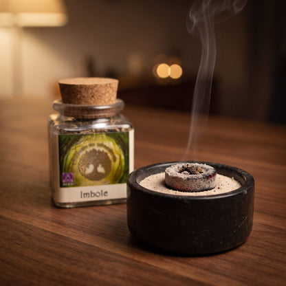 Imbolc, Jahreskreisfeste-Räuchermischung von Berk, 50 ml im Korkglas: glimmende Räucherkohle mit einer Prise Räucherwerk in einer Specksteinschale, dahinter das Korkglas, feiner Rauch steigt auf einer dunklen Holzplatte am Abend auf – Spirit of Gaia