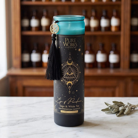 Manifestationskerze Eye of Providence (Salbei & weißer Tee) von Pure Works: hohe Glaskerze mit türkisfarbenem Glasrand, schwarzer Quaste und Goldanhänger auf Marmortheke vor Apothekerschrank, daneben getrocknete Salbeiblätter – Spirit of Gaia
