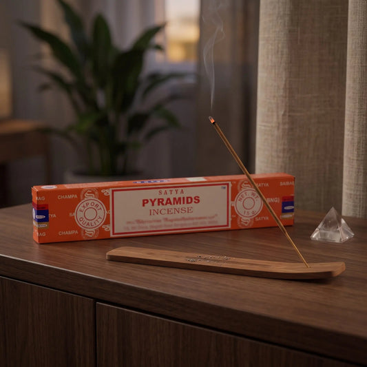 Satya Pyramids Räucherstäbchen: glimmendes Räucherstäbchen mit feinem Rauch im Holzhalter, dahinter die Packung, auf einem Sideboard am Abend