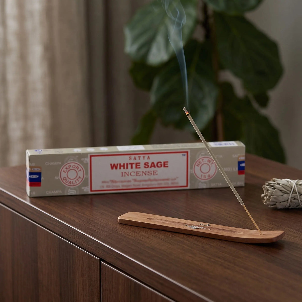 Satya White Sage Räucherstäbchen: glimmendes Räucherstäbchen mit feinem Rauch im Holzhalter, dahinter die Packung, auf einem Sideboard am Abend