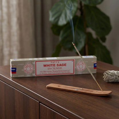 Satya White Sage Räucherstäbchen: glimmendes Räucherstäbchen mit feinem Rauch im Holzhalter, dahinter die Packung, auf einem Sideboard am Abend