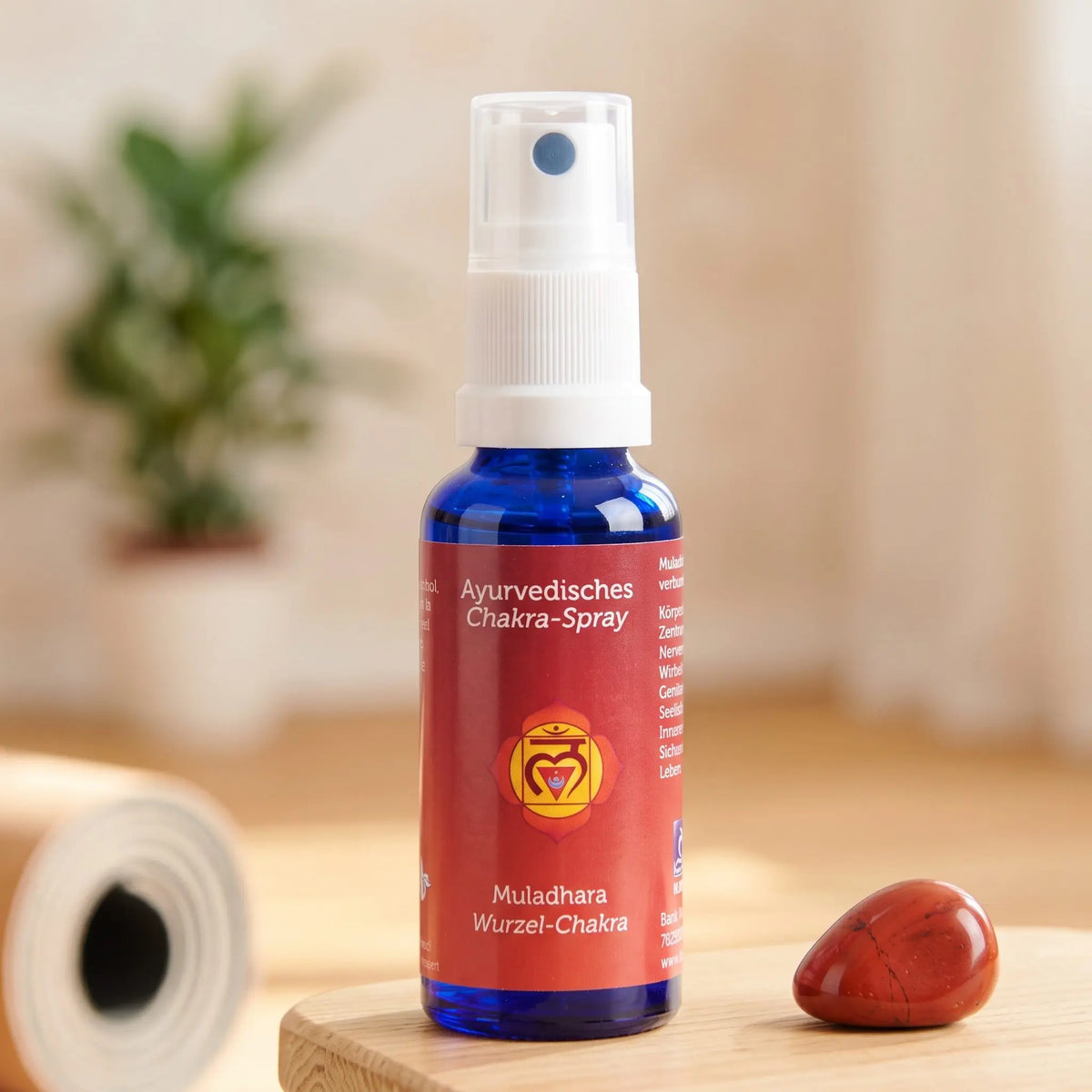 Ayurvedisches Chakra-Spray Muladhara (Wurzel-Chakra): 30-ml-Sprühflasche auf einem Holzhocker neben einer gerollten Yogamatte, daneben roter Jaspis