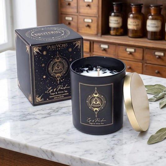 Manifestationskerze Eye of Providence mit Obsidian (Salbei & weißer Tee) von Pure Works: große Kerze im schwarzen Glas mit Golddeckel und schwarze Schachtel mit Goldprägung auf Marmortheke vor Apothekerschrank, daneben getrocknete Salbeiblätter – Spirit of Gaia