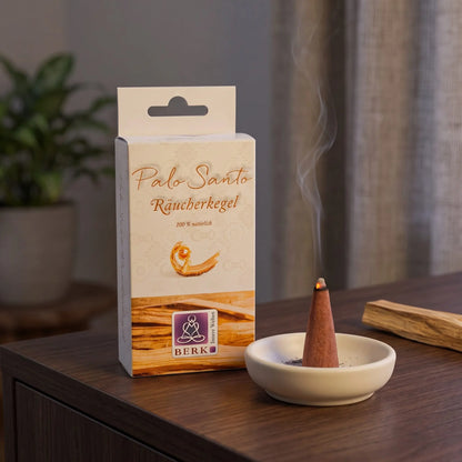 Räucherpyramiden Palo Santo von Berk: glimmender Räucherkegel mit feinem Rauch auf einem Keramiktellerchen, dahinter die Schachtel, auf einem Sideboard am Abend