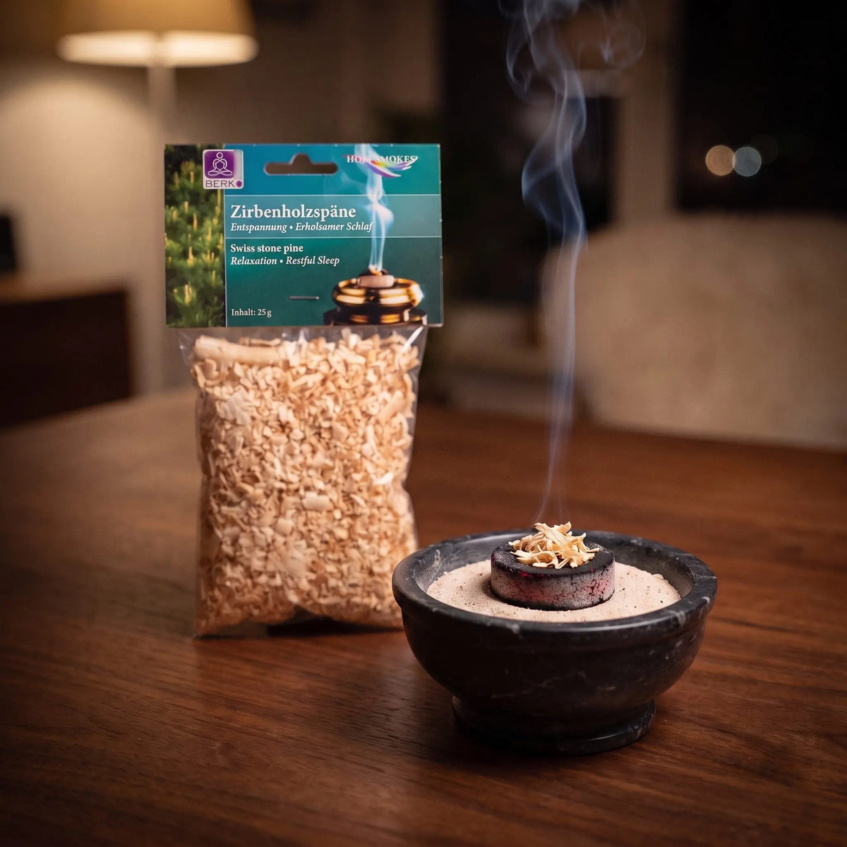 Zirbenholzspäne, sortenreines Räucherwerk von Berk, 25 g: eine Prise Zirbenholzspäne glimmt auf der Räucherkohle, dahinter die Tüte, feiner Rauch steigt auf einer dunklen Holzplatte am Abend auf – Spirit of Gaia
