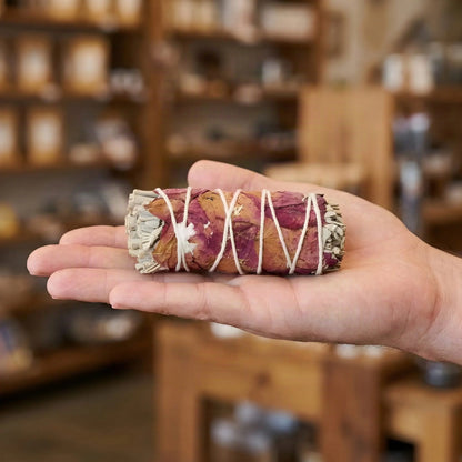 Größenvergleich: das ca. 10 cm lange Salbeibündel mit Rosenblättern liegt auf einer offenen Hand vor Ladenregalen