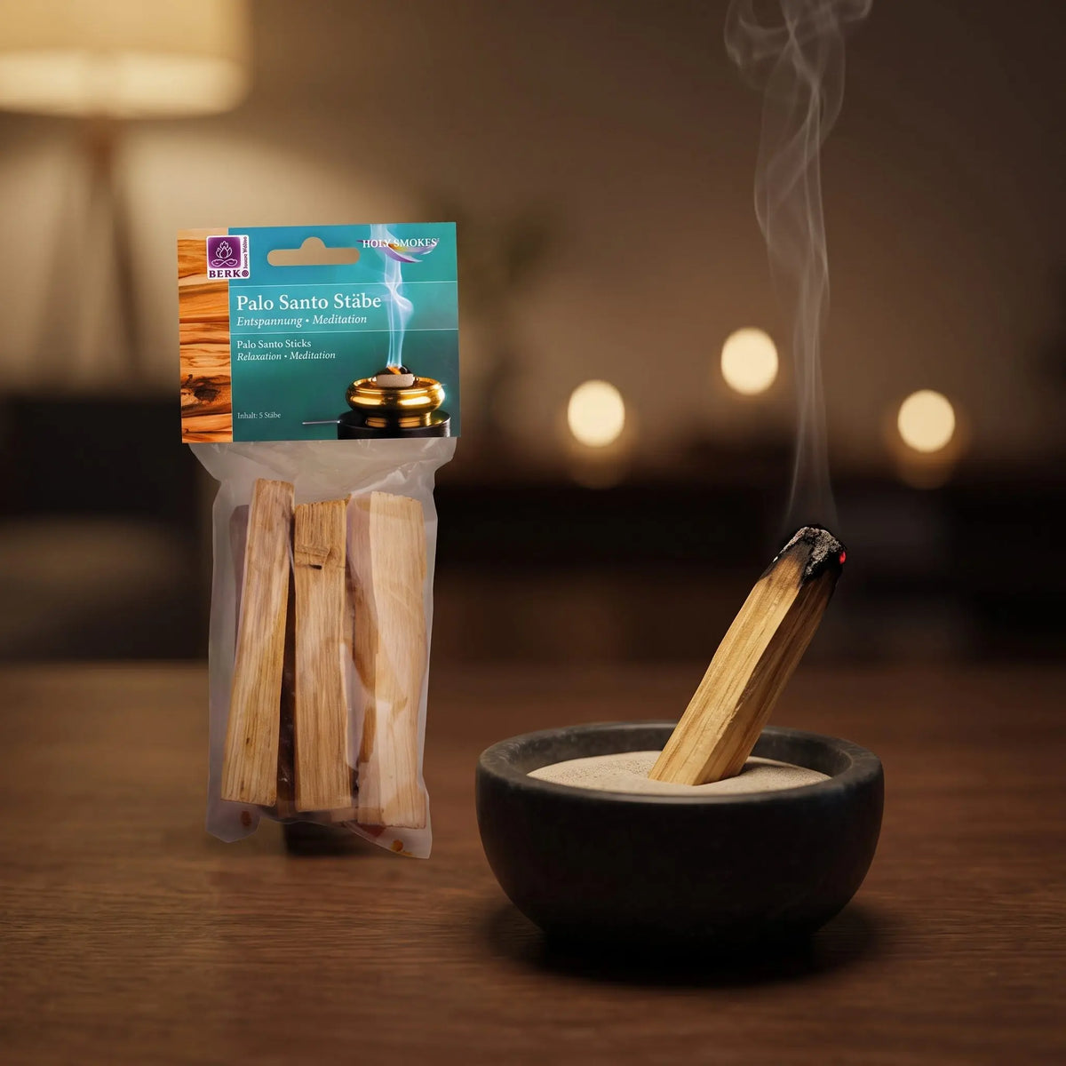Palo Santo Holzstäbe, 5 Stück, von Berk: ein Palo-Santo-Stab steckt schräg in einer Sandschale und glimmt an der Spitze, dahinter die Tüte, feiner Rauch steigt auf einer dunklen Holzplatte am Abend auf – Spirit of Gaia