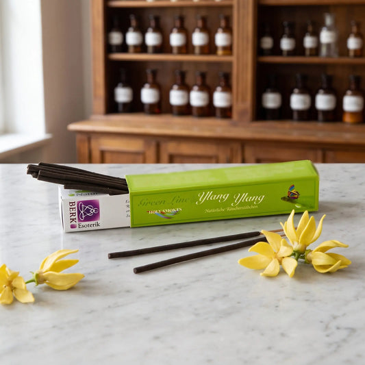 Ylang Ylang Räucherstäbchen naturrein, Holy Smokes Green Line: geöffnete Packung auf einer Marmortheke vor einem Apothekerschrank, davor zwei lose Räucherstäbchen ohne Bambuskern und Ylang-Ylang-Blüten – Spirit of Gaia
