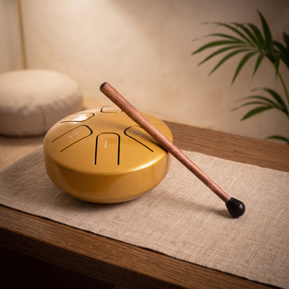 Kleine goldene Steeldrum mit Klangstab auf einem Leinenläufer, dahinter ein warmer Meditationsraum