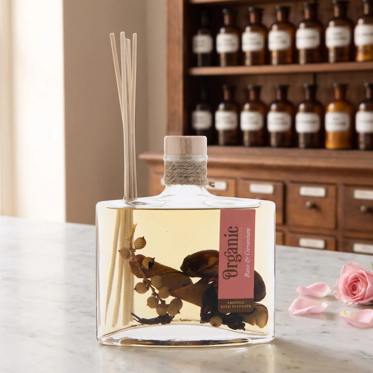 Reed Diffuser Rose und Geranie von Organic Goodness: eckige Glasflasche mit Holzstopfen, Jutekordel und Schilfrohrstäbchen auf einer Marmortheke vor einem Apothekerschrank