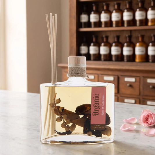 Reed Diffuser Rose und Geranie von Organic Goodness: eckige Glasflasche mit Holzstopfen, Jutekordel und Schilfrohrstäbchen auf einer Marmortheke vor einem Apothekerschrank