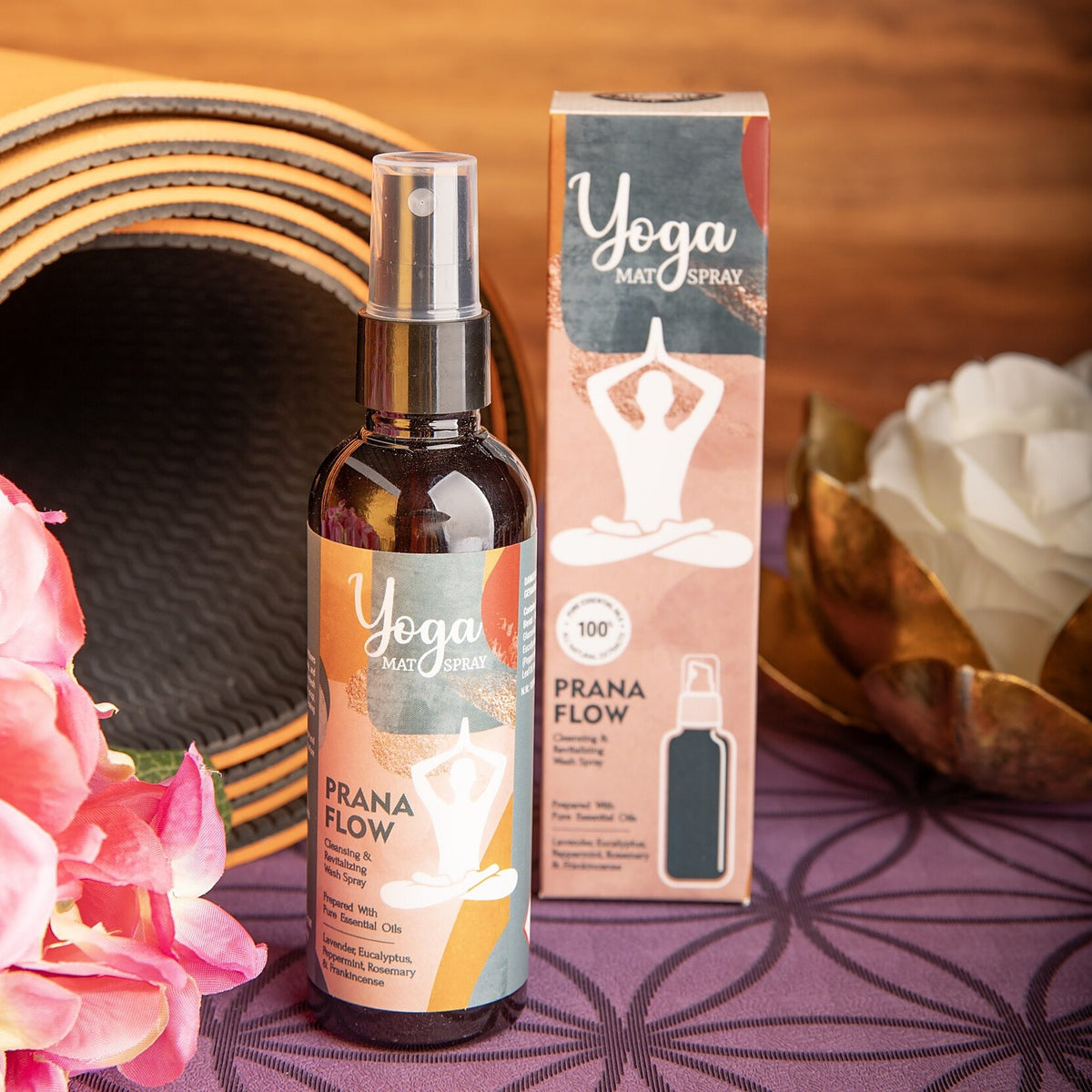 Flasche und Faltschachtel des Yogamatten-Sprays Prana Flow vor einer aufgerollten Yogamatte, rosa Blüten und einer goldenen Lotusschale auf einer lila Matte
