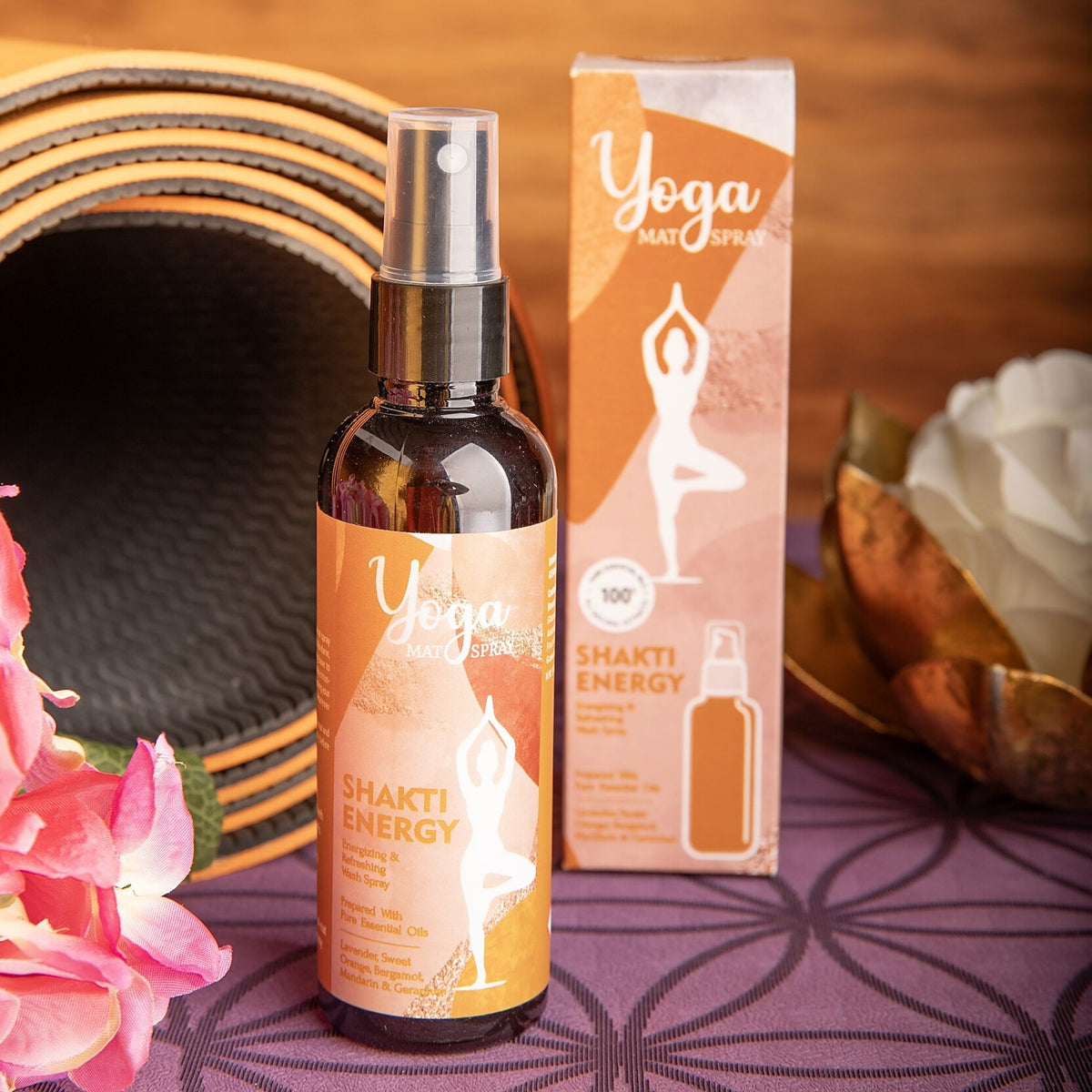 Flasche und Faltschachtel des Yogamatten-Sprays Shakti Energy vor einer aufgerollten Yogamatte, rosa Blüten und einer goldenen Lotusschale auf einer lila Matte