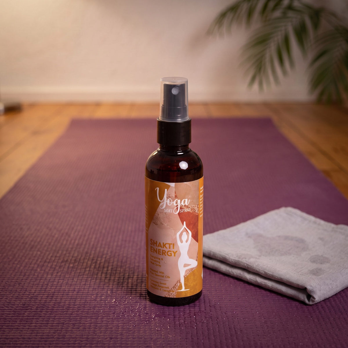 Das Yogamatten-Spray Shakti Energy steht auf einer ausgerollten lila Yogamatte, daneben ein zusammengelegtes graues Tuch, vor der Flasche feine Wassertropfen