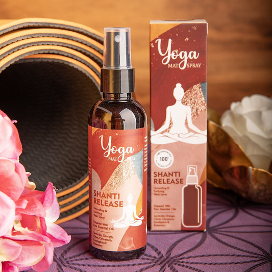 Flasche und Faltschachtel des Yogamatten-Sprays Shanti Release vor einer aufgerollten Yogamatte, rosa Blüten und einer goldenen Lotusschale auf einer lila Matte