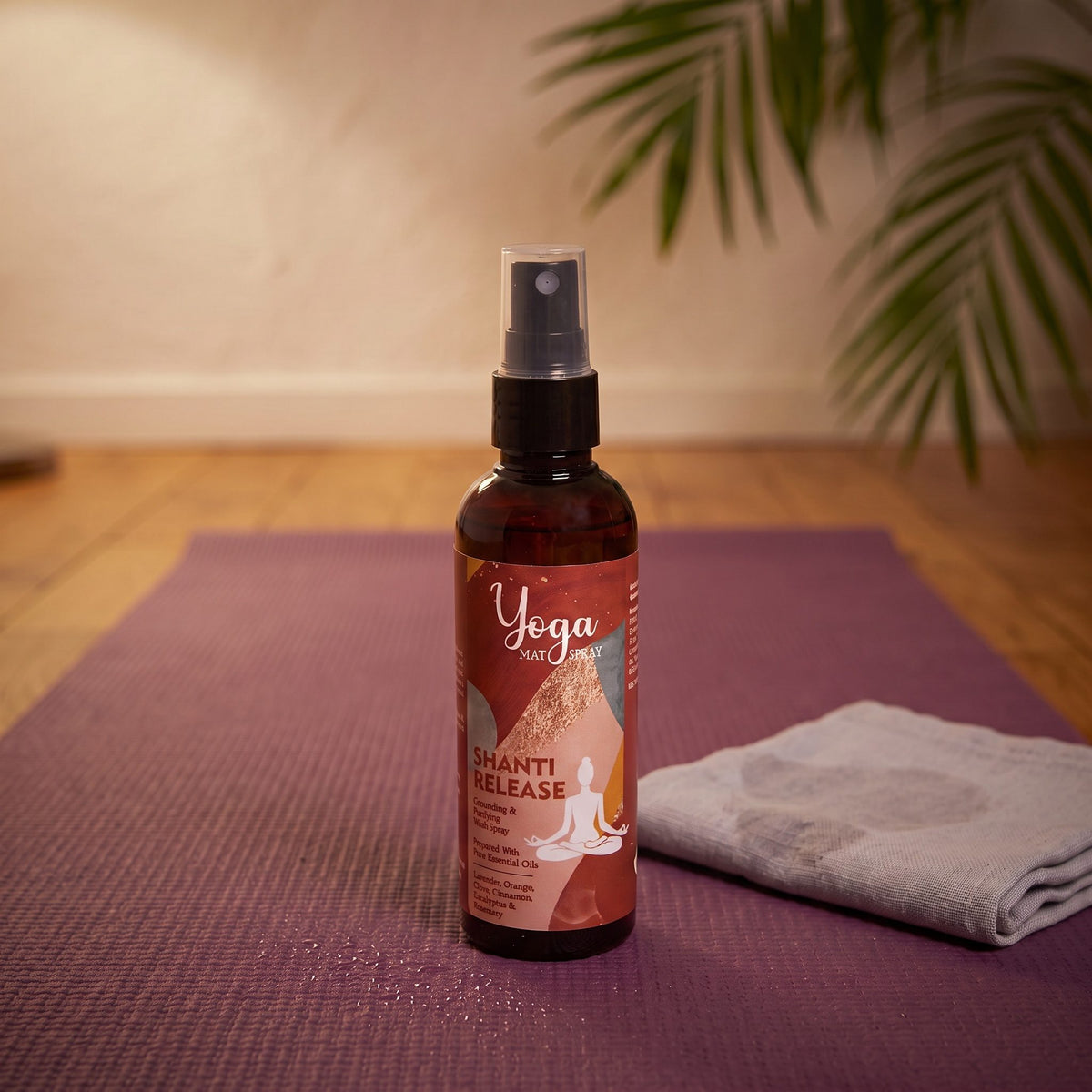 Das Yogamatten-Spray Shanti Release steht auf einer ausgerollten lila Yogamatte, daneben ein zusammengelegtes graues Tuch, vor der Flasche feine Wassertropfen