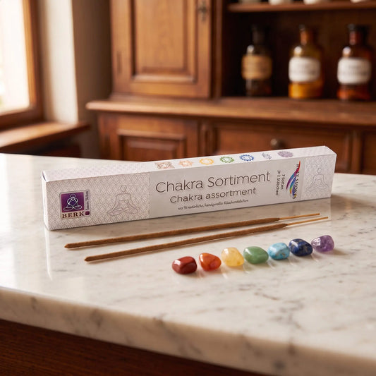 Chakra Line Sortimentspackung Holy Smokes: Packung auf einer Marmortheke vor einem Apothekerschrank, daneben zwei lose Räucherstäbchen und sieben Trommelsteine in den Chakrafarben – Spirit of Gaia